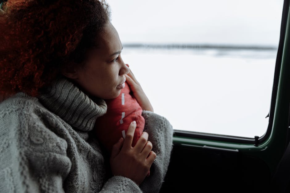 A woman with curly hair gazes thoughtfully out of a moving vehicle, holding a red pillow against a snowy backdrop.